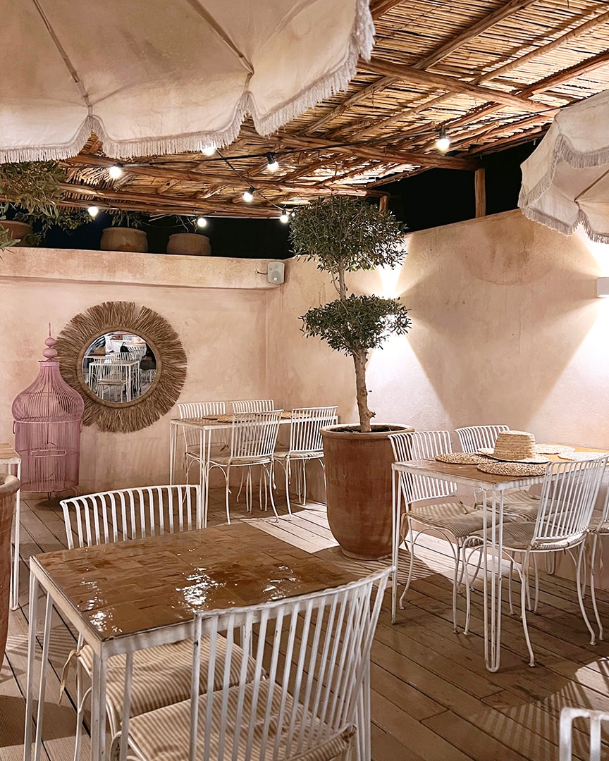 A cozy outdoor terrace with white chairs and a wooden table, under a straw canopy with small lights.