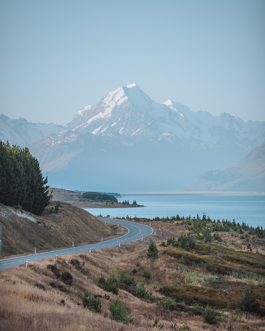 A road curving along a lakes edge with a tall snowy mountain in the background. Lake Pukaki, New Zealand South Island.