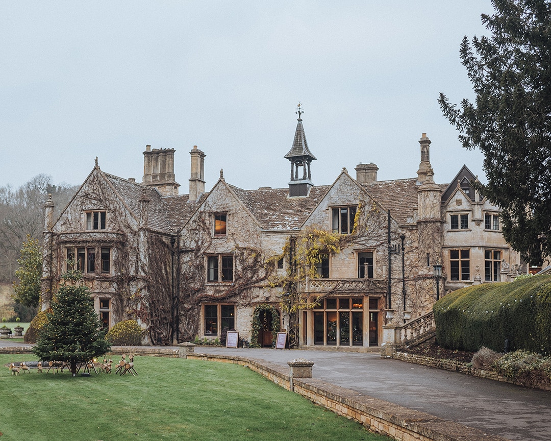 A large ivy-covered stone manor with a bell tower, gardens, and Christmas decorations outside the entrance.