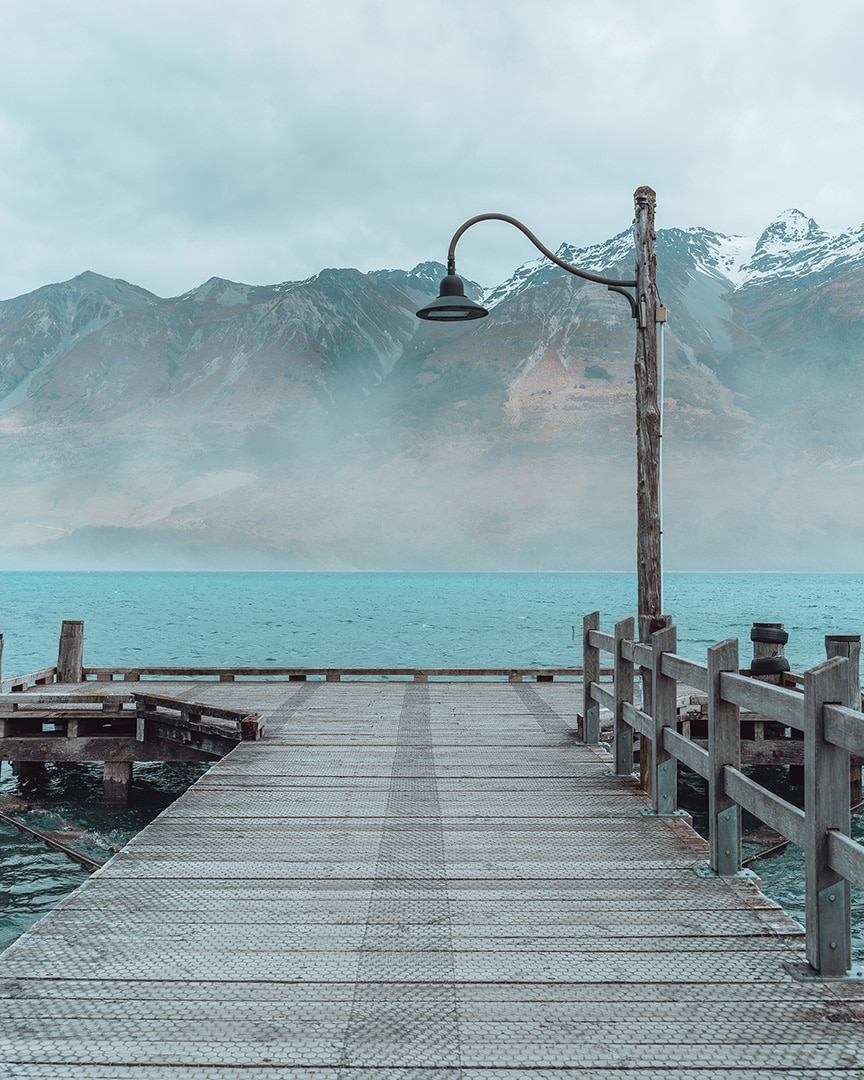 A tranquil wooden pier extends into a mist-covered lake with mountains in the background. Glenorchy, New Zealand.