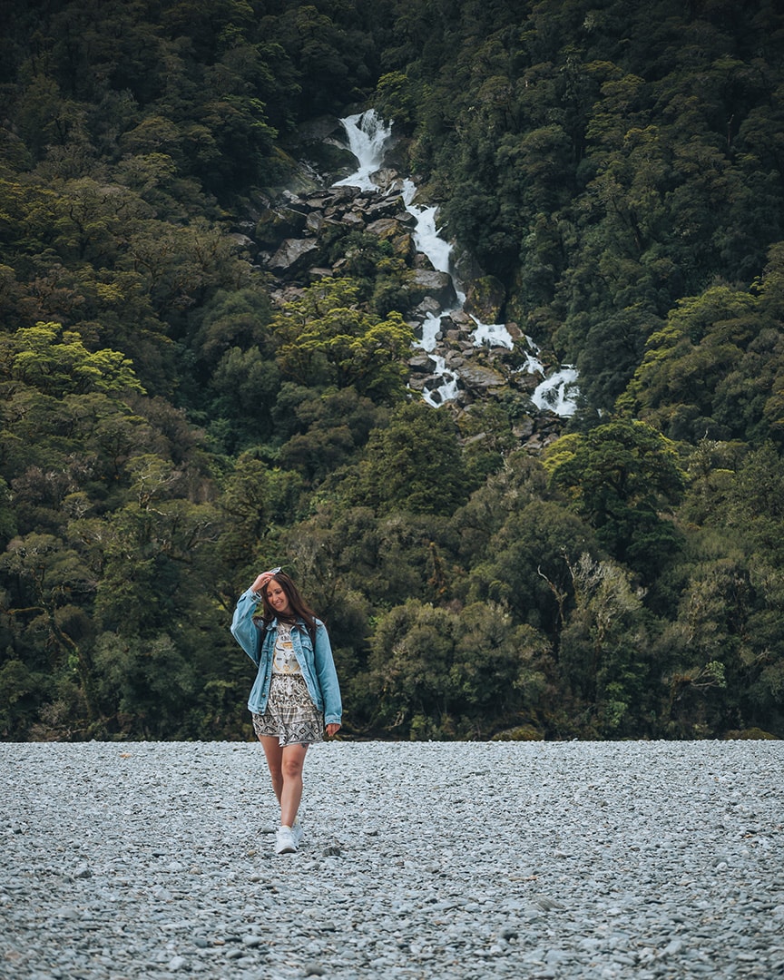 A girl standing on grey rocky ground, behind her is a lush green forest with a waterfall pouring down. Haast Pass, New Zealand.