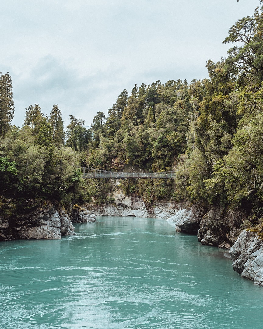 A lush green river gorge with turquoise waters and a swing bridge overhead.