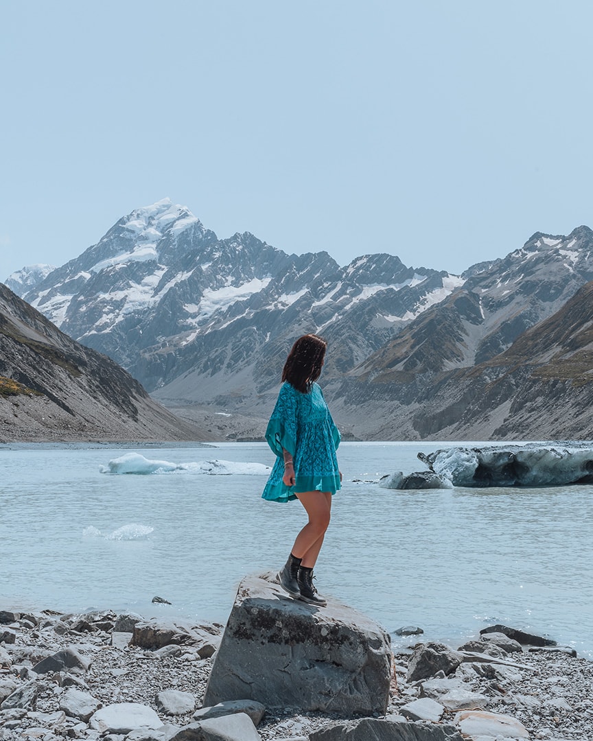 A person in a flowy blue dress stands on a rock in a glacial valley, facing a snow-capped mountain. Hooker Lake, New Zealand South Island.