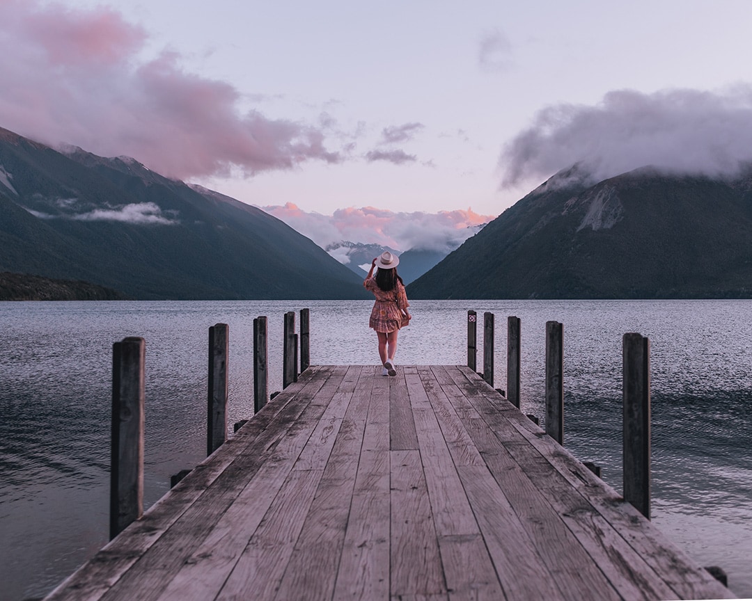 A person in a wide-brimmed hat stands at the end of a rustic wooden jetty, gazing towards a dramatic mountainous landscape under a dusky sky. The view from Kerr Bay Camping, Lake Rotoiti, South Island.