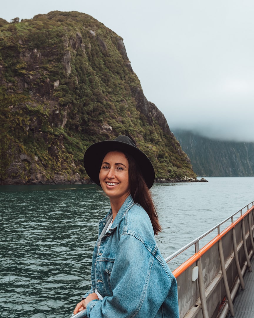 A girl leans on the railing of a boat with steep, green cliffs in the background.