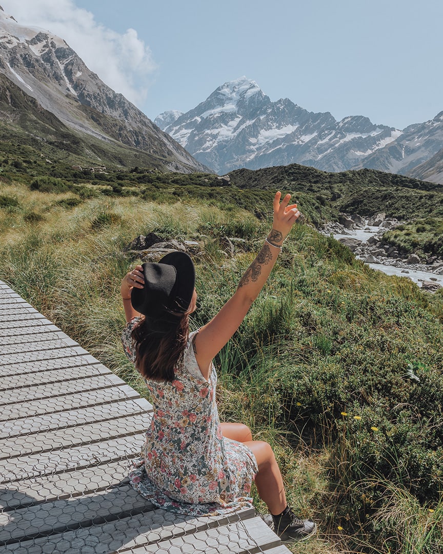 A person in a floral dress and hat sits on a wooden path, raising their arms towards a majestic snow-capped mountain in the distance. Mount Cook, one of the best places to visit in New Zealand South Island.