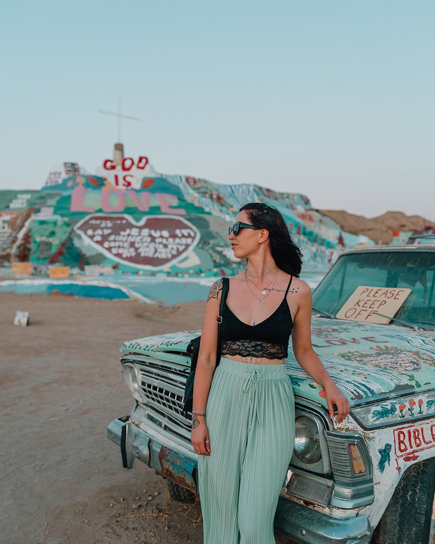 A girl in a black top and green trousers leaning on a blue car with flowers and bible verses written on it. Behind her is a man made multi colored mountain, more verses are written on it. Salvation Mountain, California