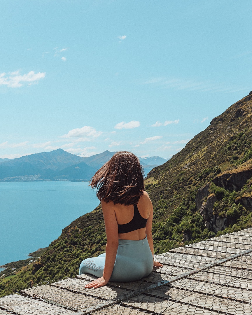 A woman sits on a wooden platform on a hill edge, with a blue lake and mountainous landscape ahead. Wye Creek, Queenstown.
