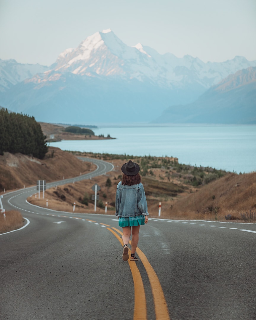 A girl walking down a road next to a turquoise lake. At the top of the lake is a tall snowy mountain. Lake Pukaki & Mount Cook, NZ South Island.