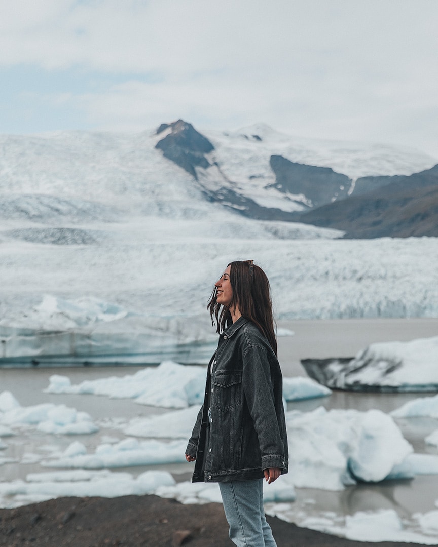 A girl wearing a black jacket standing in front of a lagoon filled with icebergs. Behind the lagoon is a mountain with a white and blue glacier.