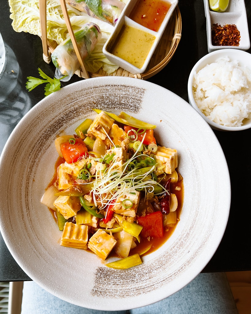 A white bowl filled with colourful green, yellow and red vegetables, tofi and a red curry sauce. 