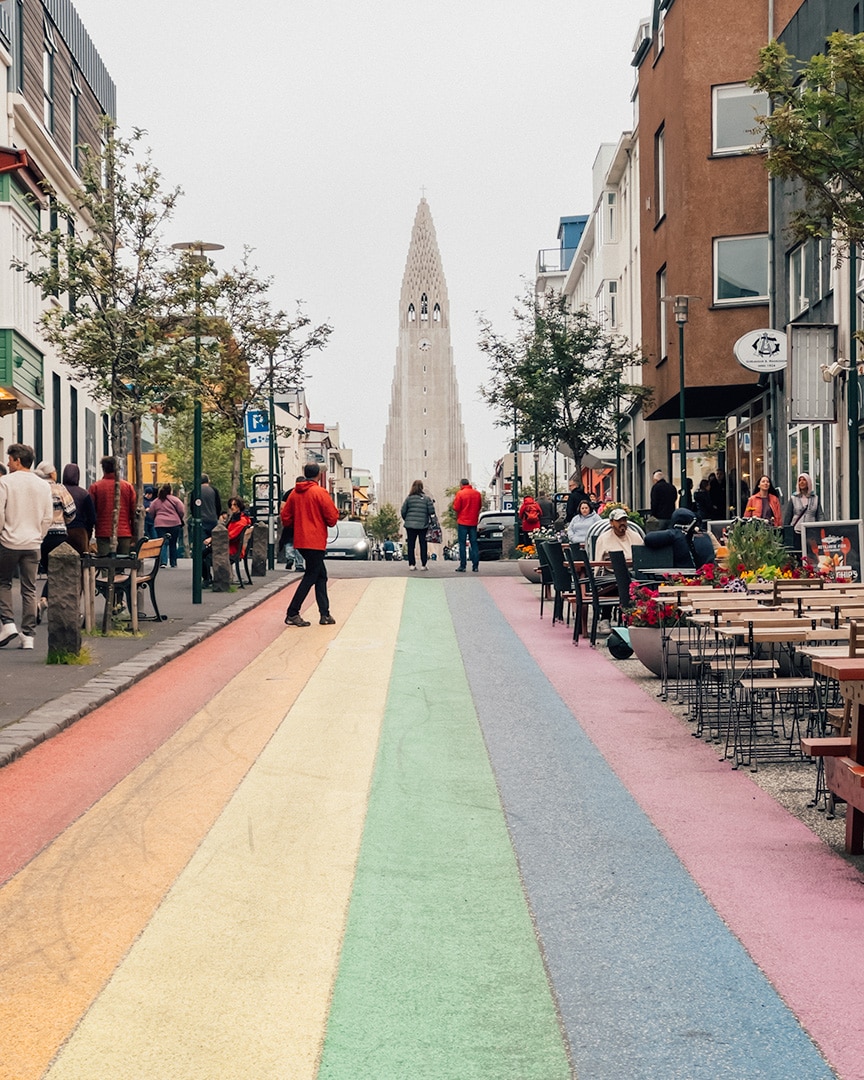 A vibrant street scene with a multicolored path that resembles a rainbow stretching down the middle of the road. This pathway draws the eye straight towards a tall church with a pointed, tower-like structure in the background. 