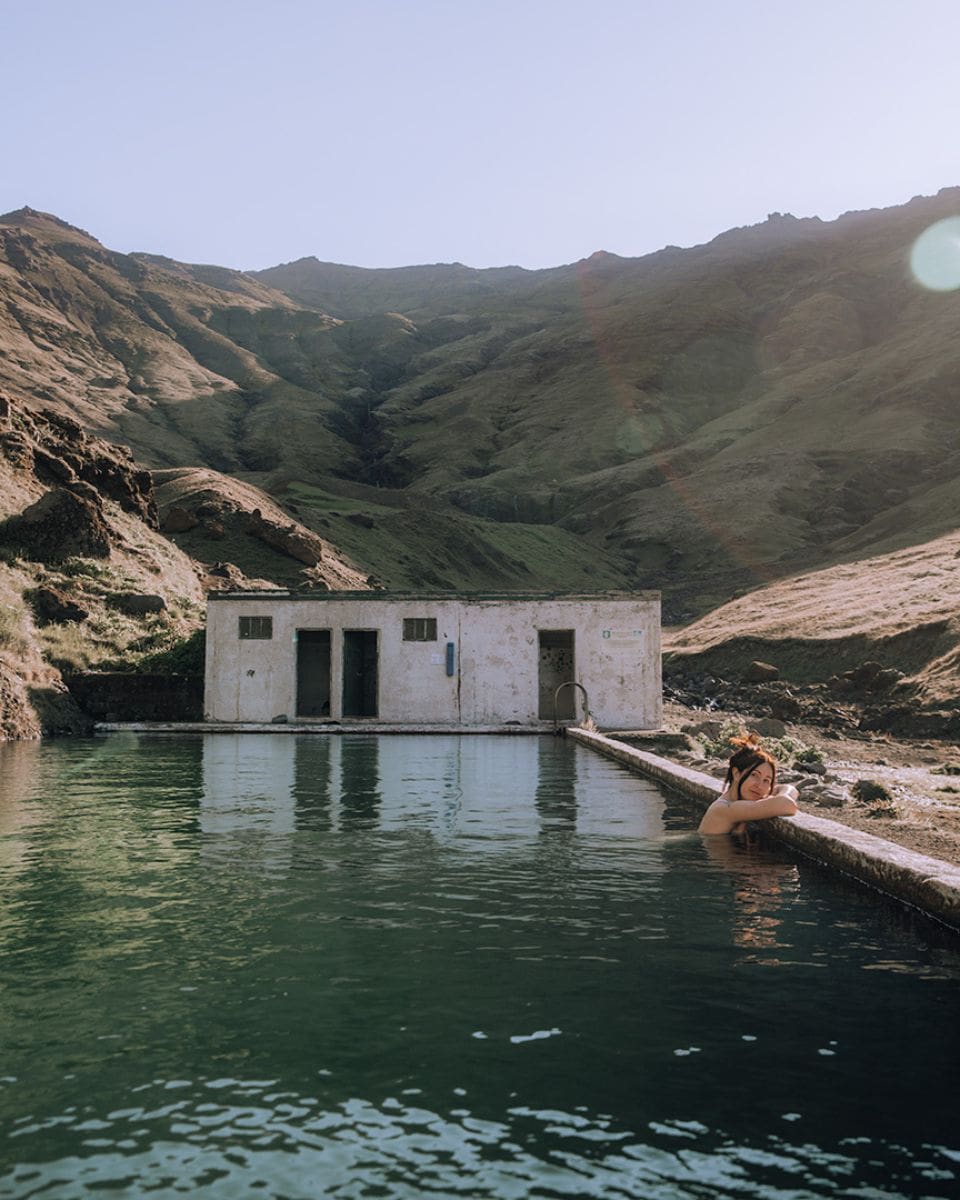 A woman relaxes in a remote outdoor swimming pool, leaning on the edge with her arms resting on the side. The water is deep green, and the pool sits in a wide, open valley surrounded by tall, grassy mountains. Behind her, there’s a small, weathered white building with changing rooms.