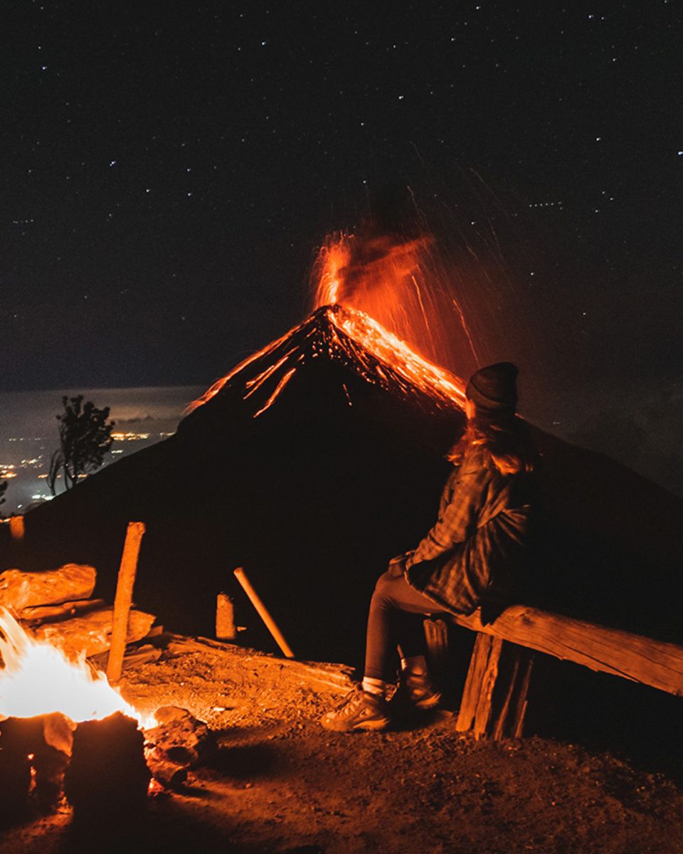 At night, a woman sits on a wooden bench near a glowing campfire, watching an active volcano erupt in the distance. Lava pours down the sides of the volcano and bursts into the sky, lighting up the darkness with red and orange streaks. She’s bundled up in a beanie and warm clothes, with the fire casting a warm glow on her face. The night sky above is filled with stars.