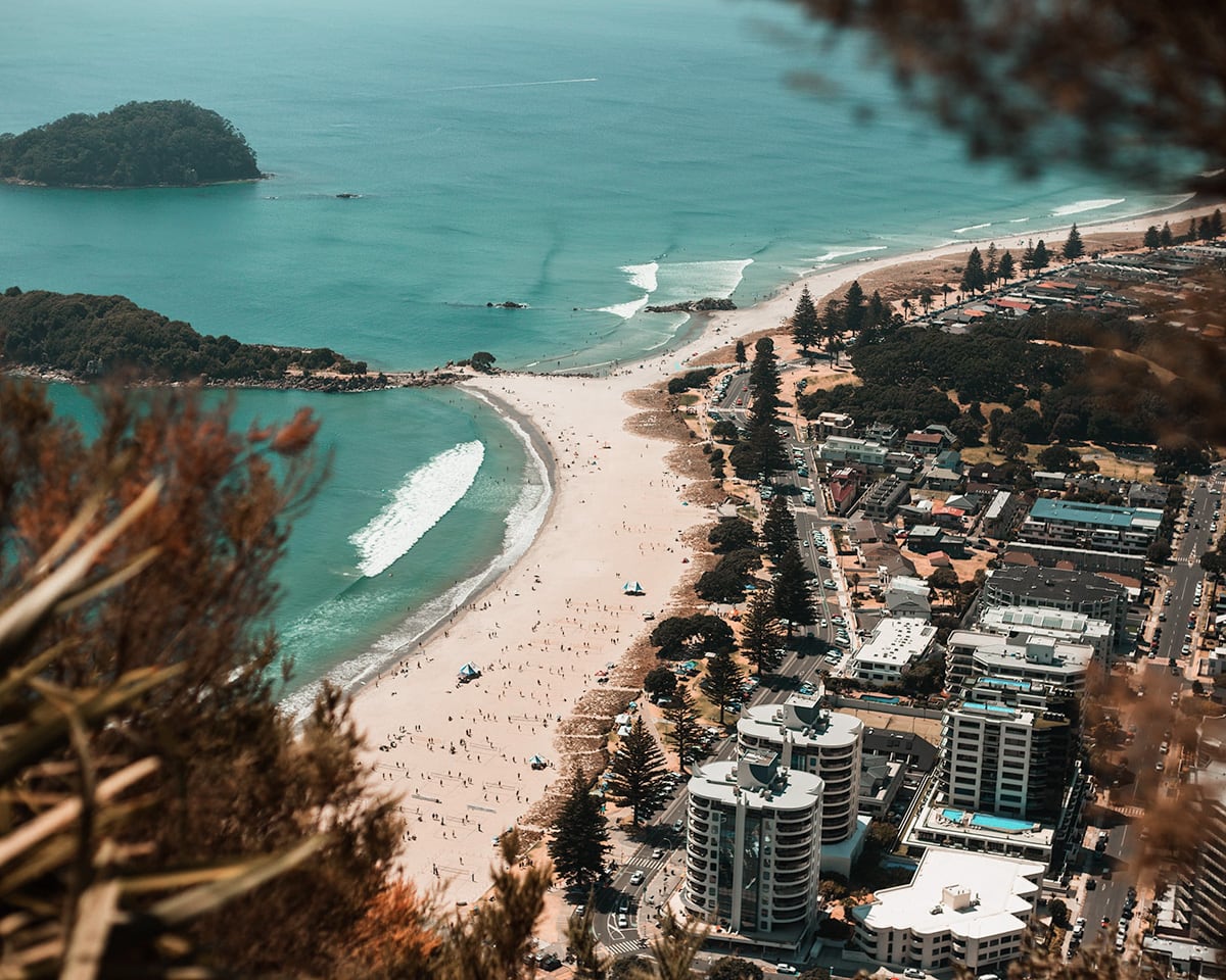 A view from above Mount Maunganui, looking down at a curved beach with people and surfers, adjacent to a calm bay. In the background is a small, forested island. The nearby urban area includes buildings, streets, and parked cars, all in close proximity to the sandy beach.