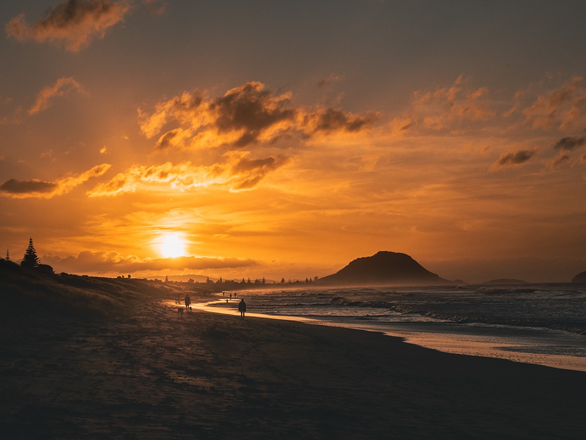 A sunset at the beach with the silhouette of Mount Maunganui in the distance. The sky is ablaze with orange and golden hues, reflecting on the waves of the ocean. People can be seen along the shoreline. The beach stretches toward the mountain.