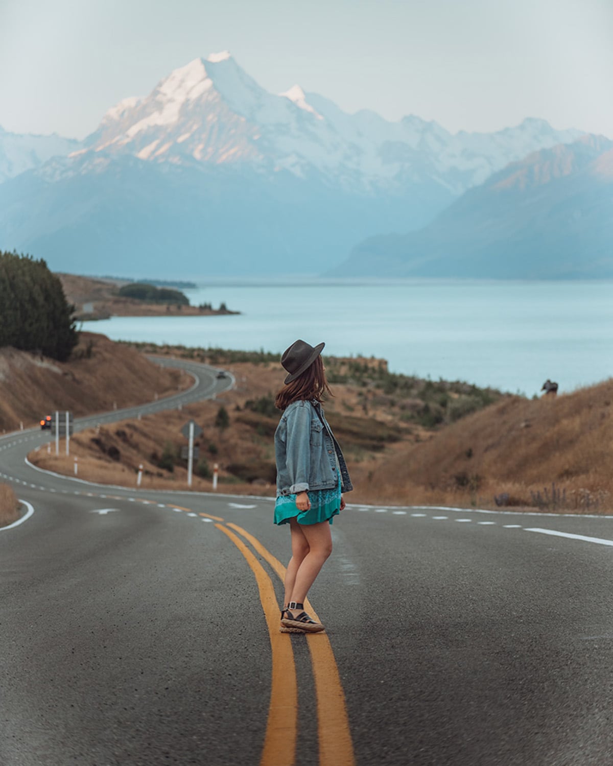 A girl stands in the middle of a winding road, facing a large, snow-capped mountain in the distance. The mountain looms over a calm lake to the left, with gentle slopes leading down to the water. The traveler is wearing a wide-brimmed hat, a denim jacket, and a turquoise skirt.