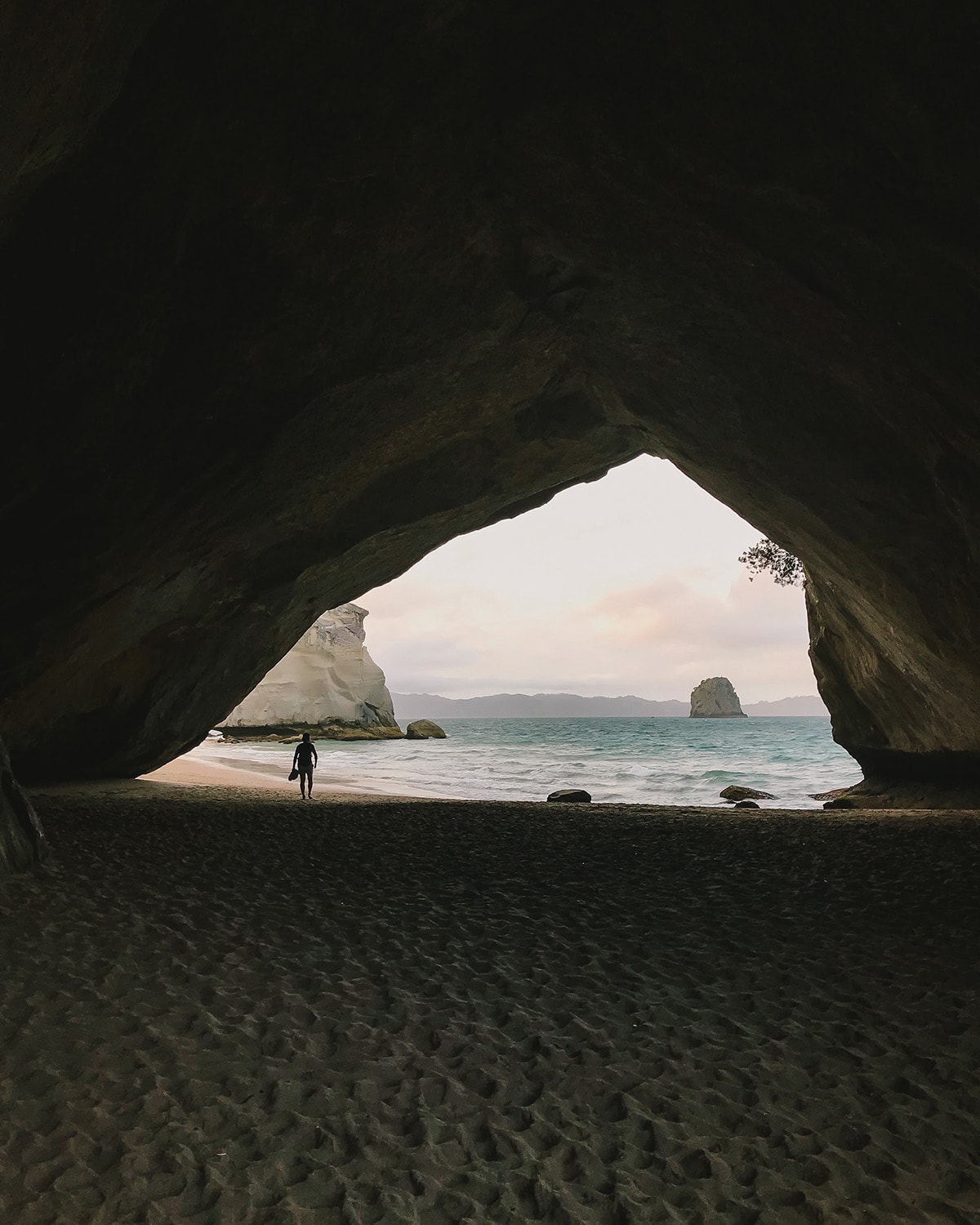 A silhouette of a person is framed by the large opening of a sea cave, with a view of the ocean and distant rock formations visible through the archway. The cave's interior shows textured walls and a sandy floor, leading out to a beach where the waves gently lap at the shore.