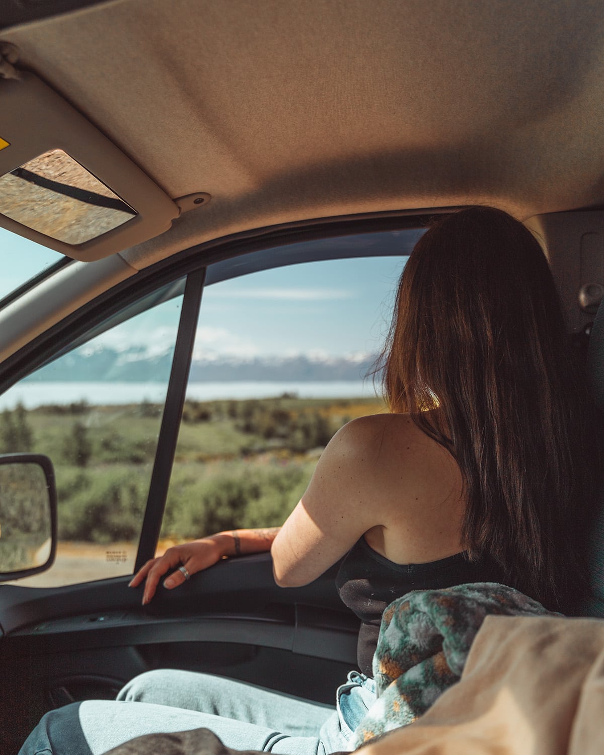 Inside a vehicle, a woman gazes out the window towards a distant mountain range. Iceland where many locals speak English.