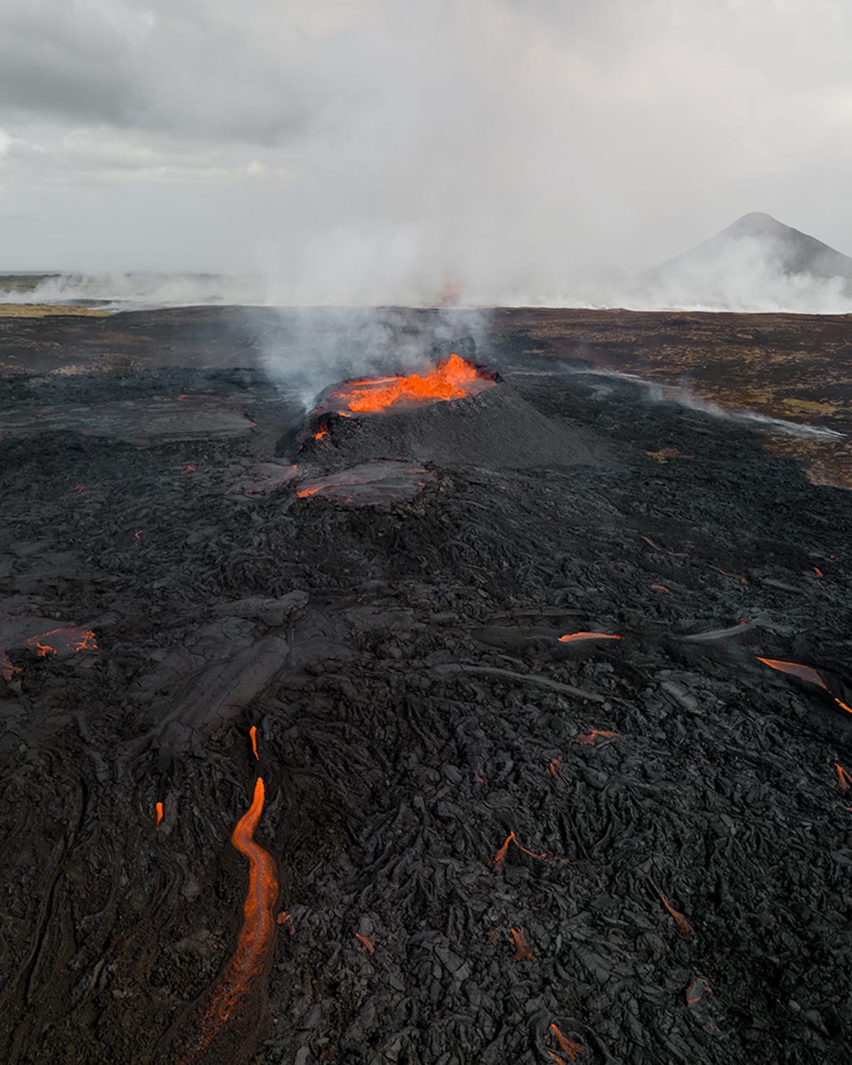 A live volcano spewing bright orange lava. Around the volcano is hardened black lava with orange and black patterns flowing away from it