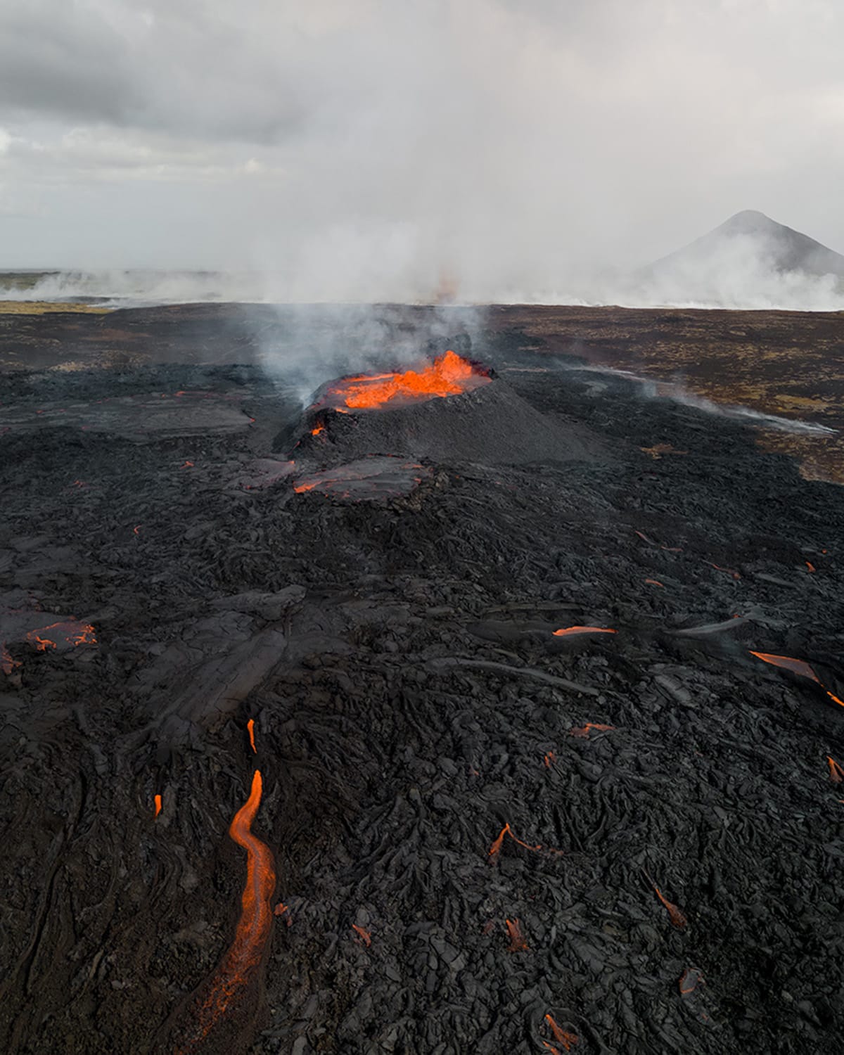 An active volcanic vent in Iceland's Reykjanes Peninsula emits smoke and flames, with molten lava snaking across the charred landscape. In the background, a volcanic cone rises against a hazy sky. The foreground is dominated by cooled, textured lava flows.