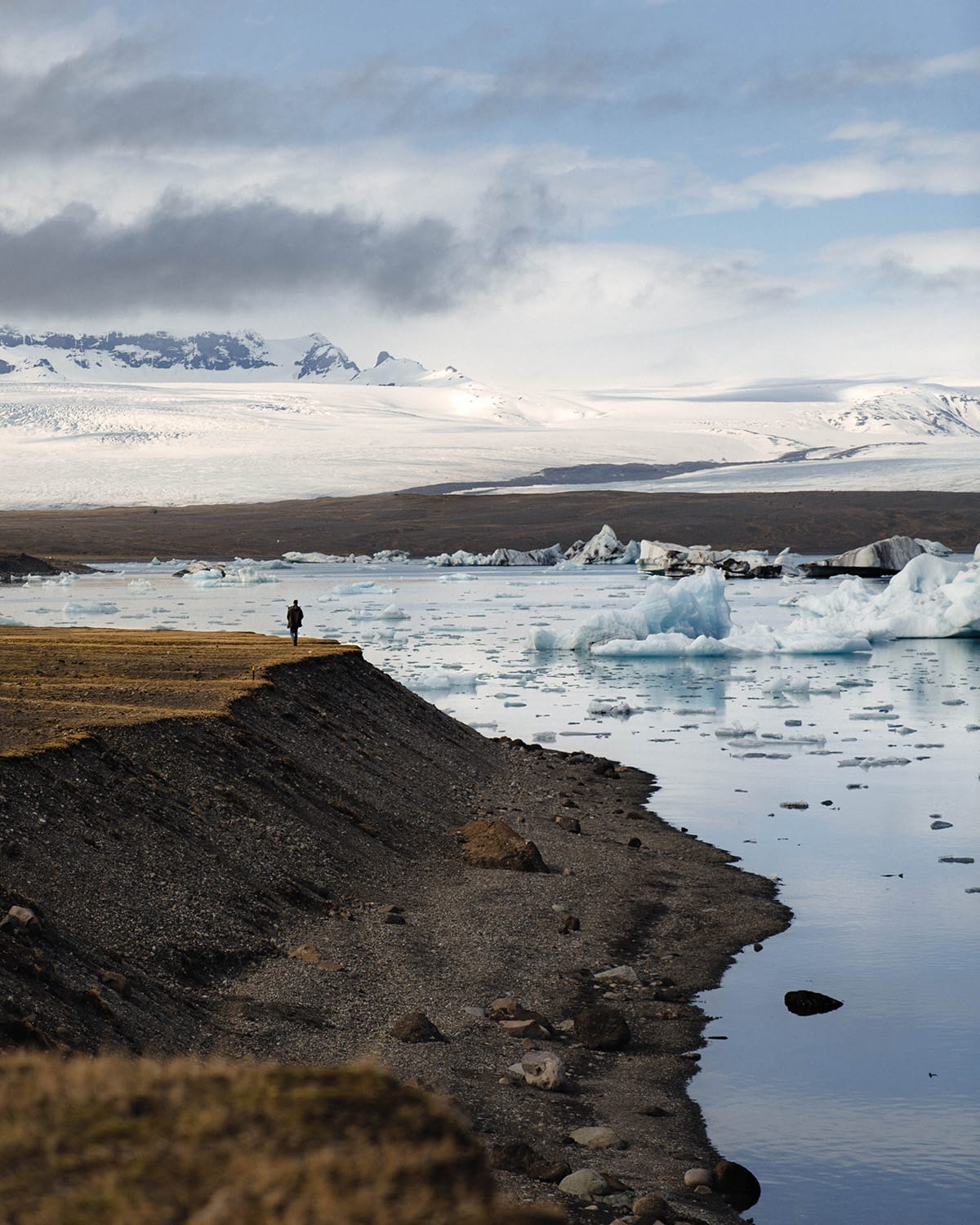 A solitary figure stands at the edge of Jökulsárlón Glacier Lagoon in Iceland, gazing out over the water dotted with icebergs. The glacier in the background extends into the mountains under a sky with light cloud cover. The shore curves gently into the lagoon.