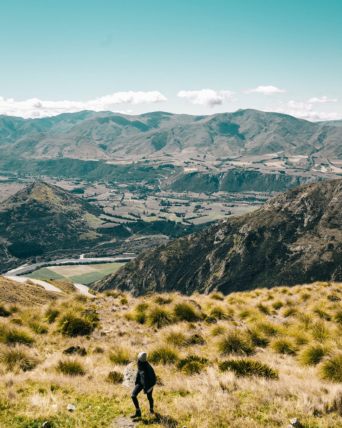 A woman stands on a hillside, gazing out at the expansive view of the Remarkables mountain range in New Zealand. The tussock-covered terrain in the foreground slopes down to a tapestry of valleys and peaks, with winding rivers and patchwork fields visible in the distance.