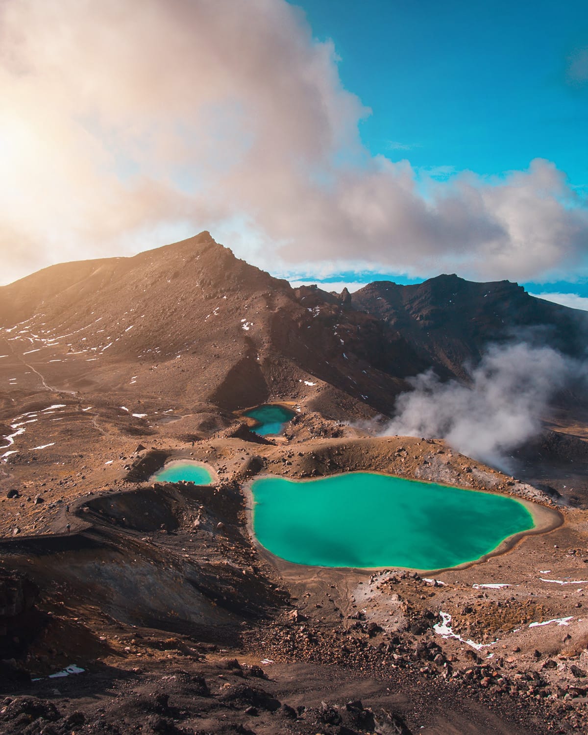 The vibrant emerald lakes of Tongariro Alpine Crossing are nestled within a stark volcanic landscape under a warm, glowing sky. Wisps of steam rise from the rugged terrain, emphasizing the area's geothermal activity. This otherworldly scene is a part of one of the most famous day hikes in New Zealand and a bucket list destination for avid hikers.