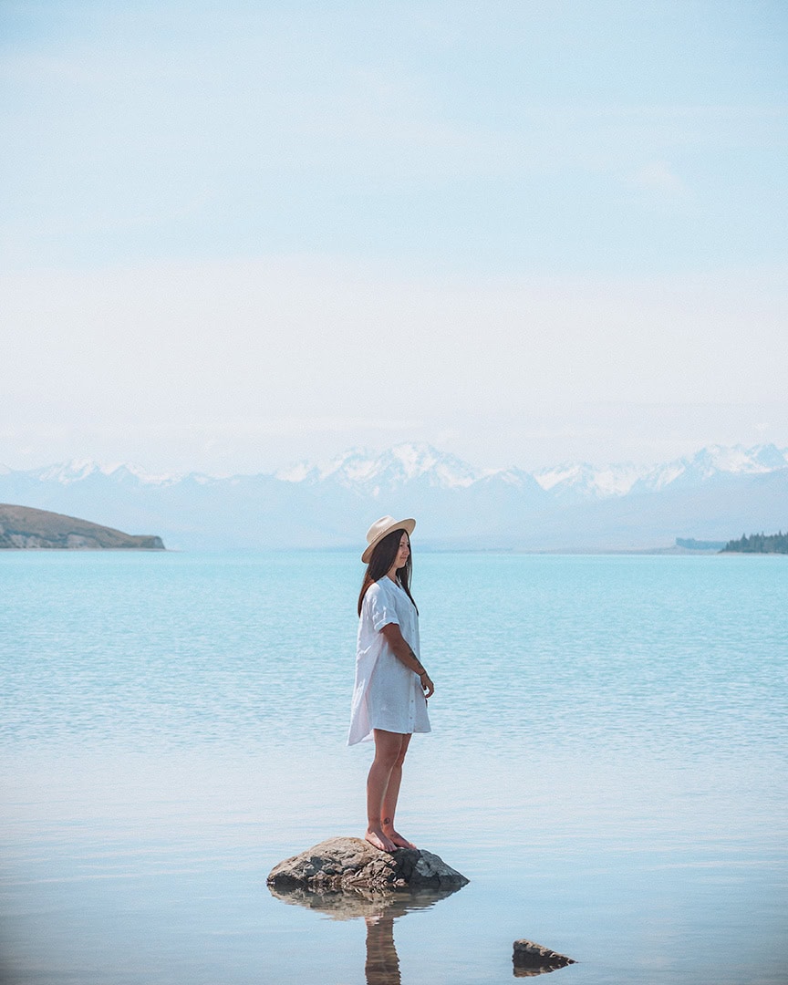 A person in a white shirt stands on a rock in a clear blue lake with snowy mountains in the distance. Lake Tekapo.