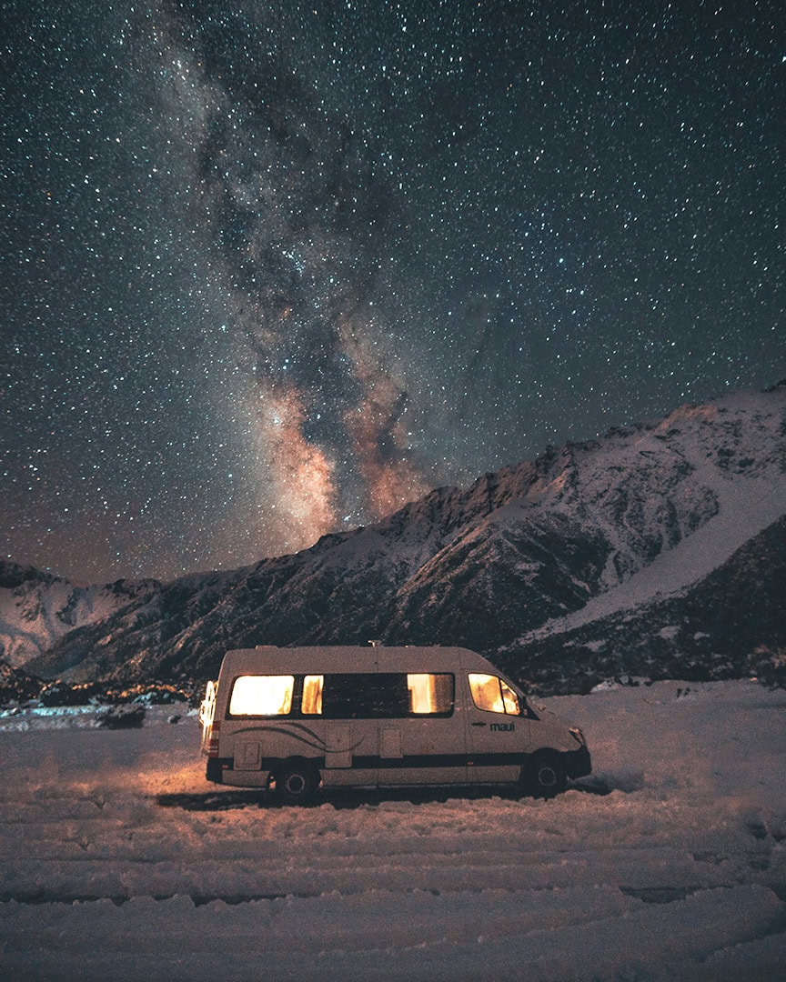 Under a star-filled sky, a camper van is illuminated from within against a backdrop of Mount Cook.
