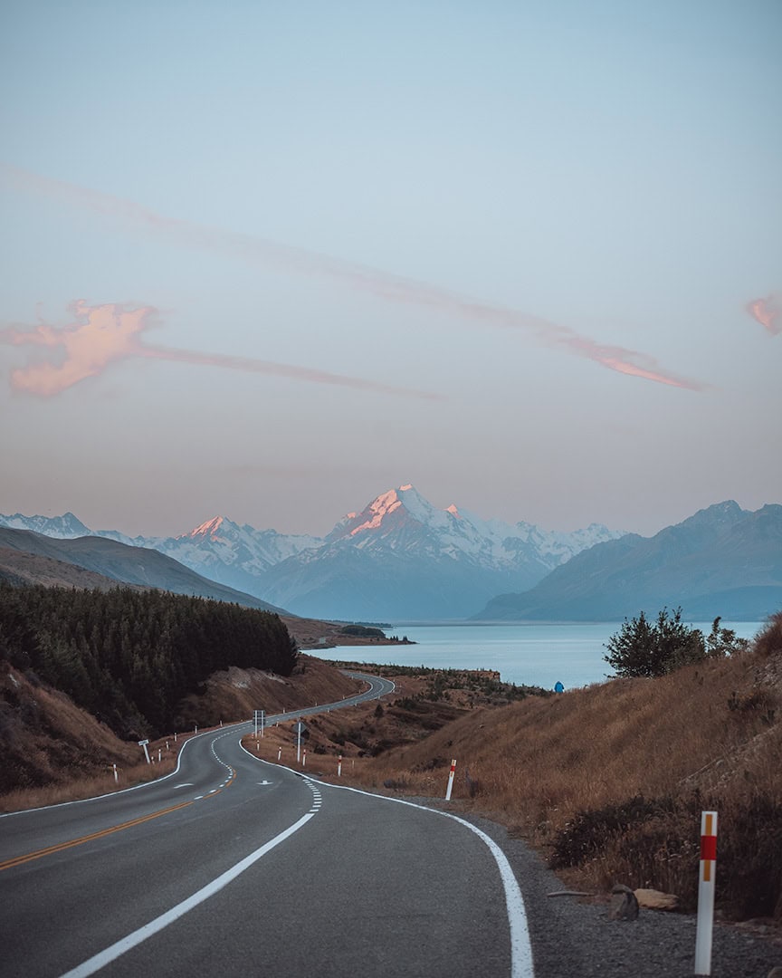 A winding road leads towards the majestic, snow-capped Mount Cook, with vibrant streaks of sunset in the sky. The road to Glentanner camping, New Zealand South Island.