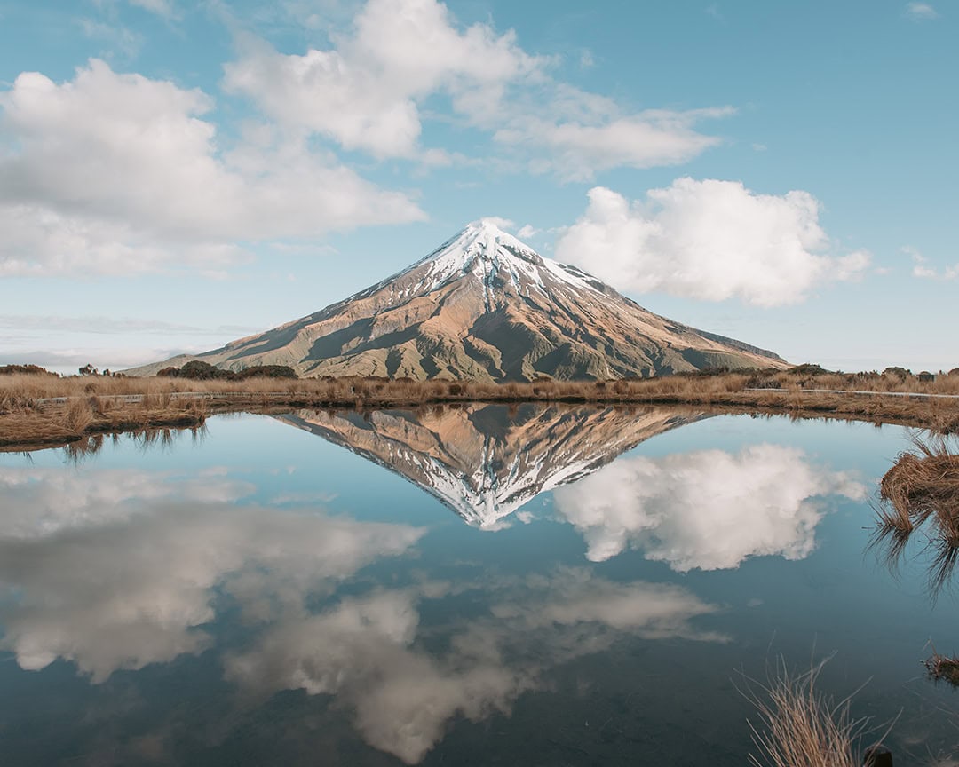 A snow-capped Mount Taranaki is perfectly reflected in a calm lake under a blue sky with scattered clouds.
