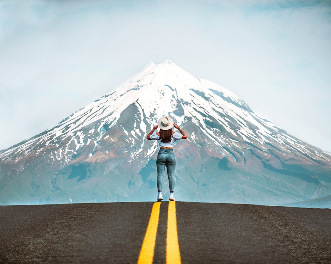 A person stands in the middle of a road, facing Mount Taranaki in the distance, wearing a hat and looking at the snow-capped mountain.