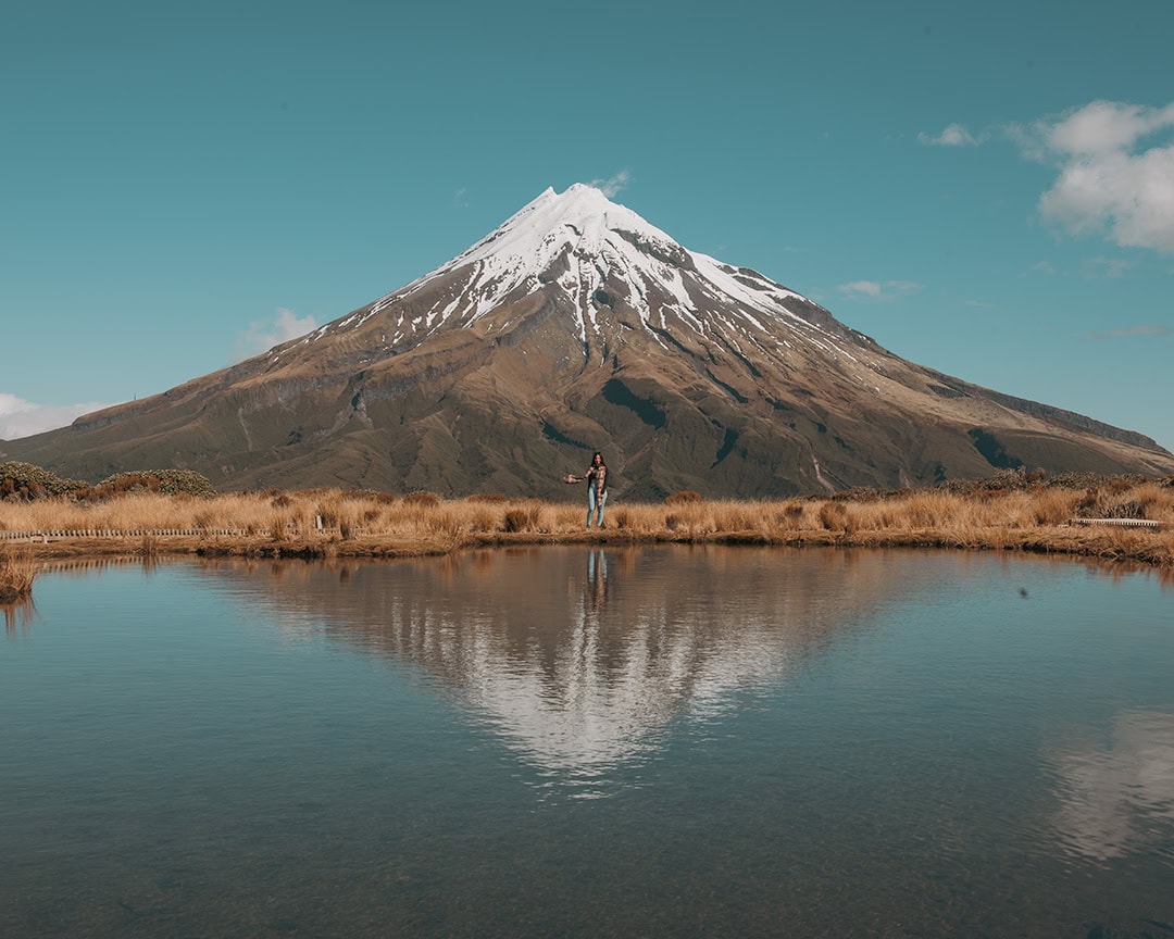 A person stands near the edge of a lake at the Pouakai Tarns walk, with Mount Taranaki and its reflection in the water, under a clear blue sky.