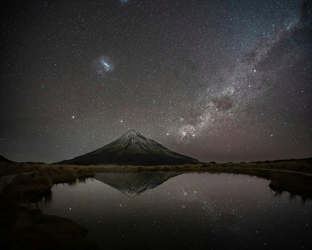 Mount Taranaki is seen at night with a starry sky above, reflected in a calm lake.