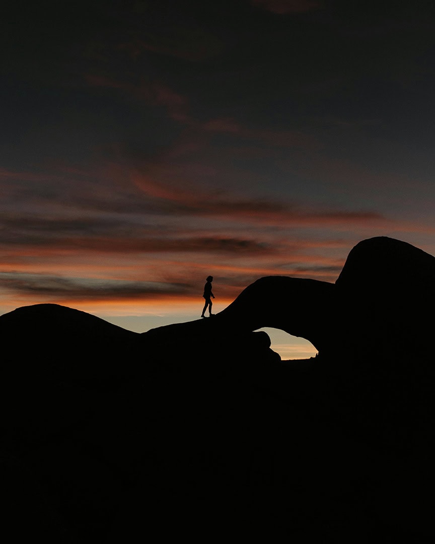 A person walking along a natural rock arch silhouetted against a colorful sunset sky.