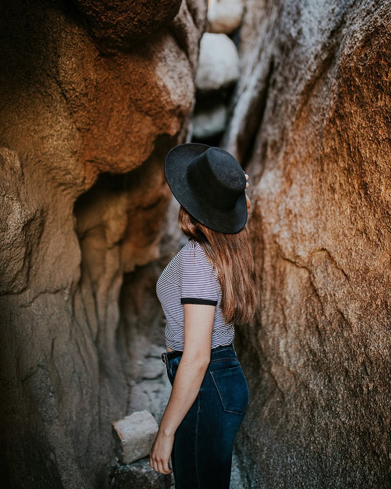 A woman with long brown hair wearing a hat exploring a narrow rock passageway.