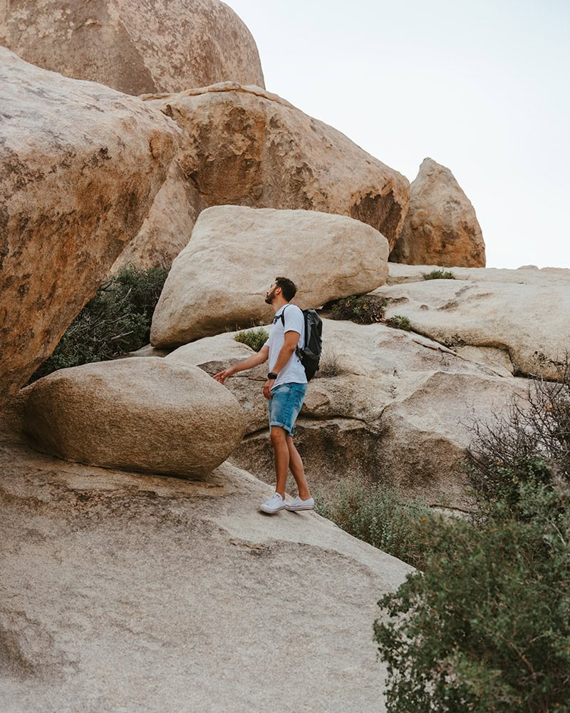 A man with a backpack climbing on a rocky formation in a desert area in Joshua Tree National Park.