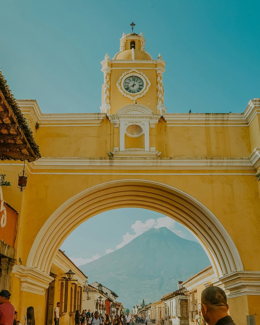 A bright yellow arch with a clock tower in Antigua, Guatemala. A volcano rises in the background under a clear blue sky, and people are walking through the cobblestone street below.