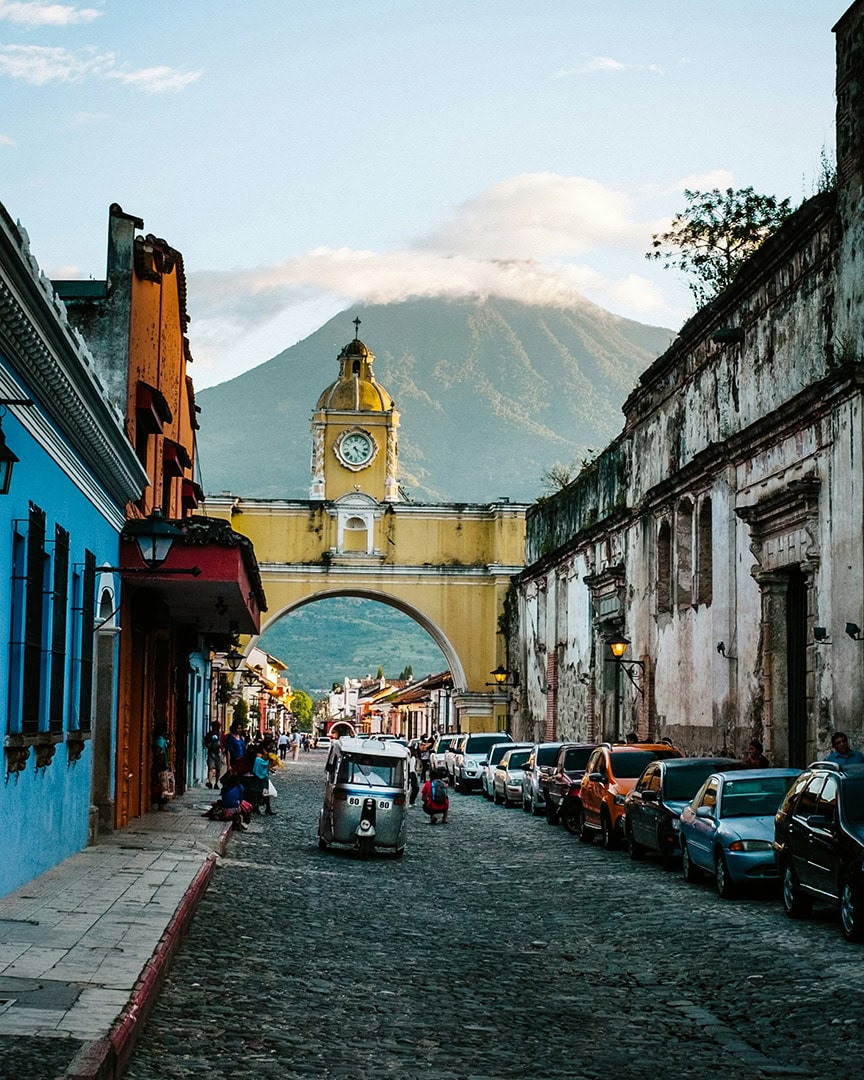 A cobblestone street lined with colorful buildings leads to a bright yellow clock tower, with a volcano rising in the background. First stop on this Guatemala 10 day itinerary.