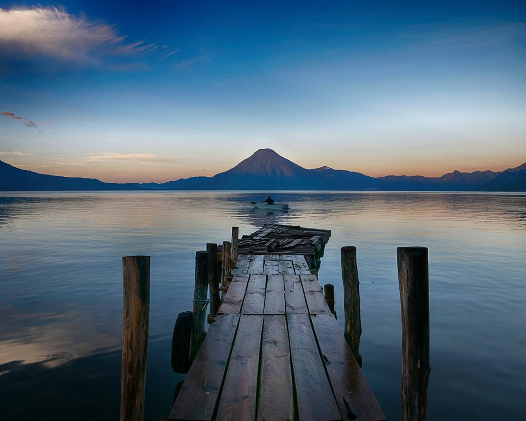 A rustic wooden dock extends into a calm lake, with a volcano silhouetted against the fading light of sunset. Lake Atitlán