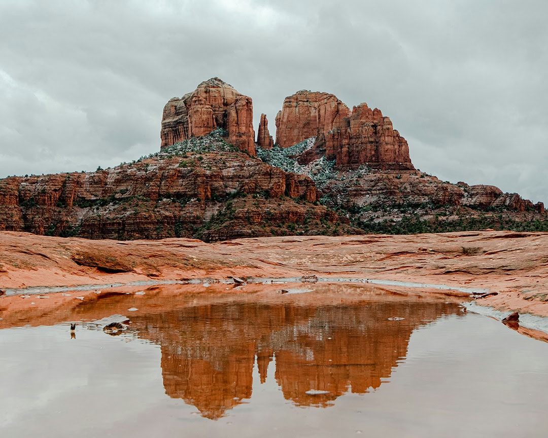 A large red rock formation with patches of snow, reflected in a puddle on the ground. Sedona in winter