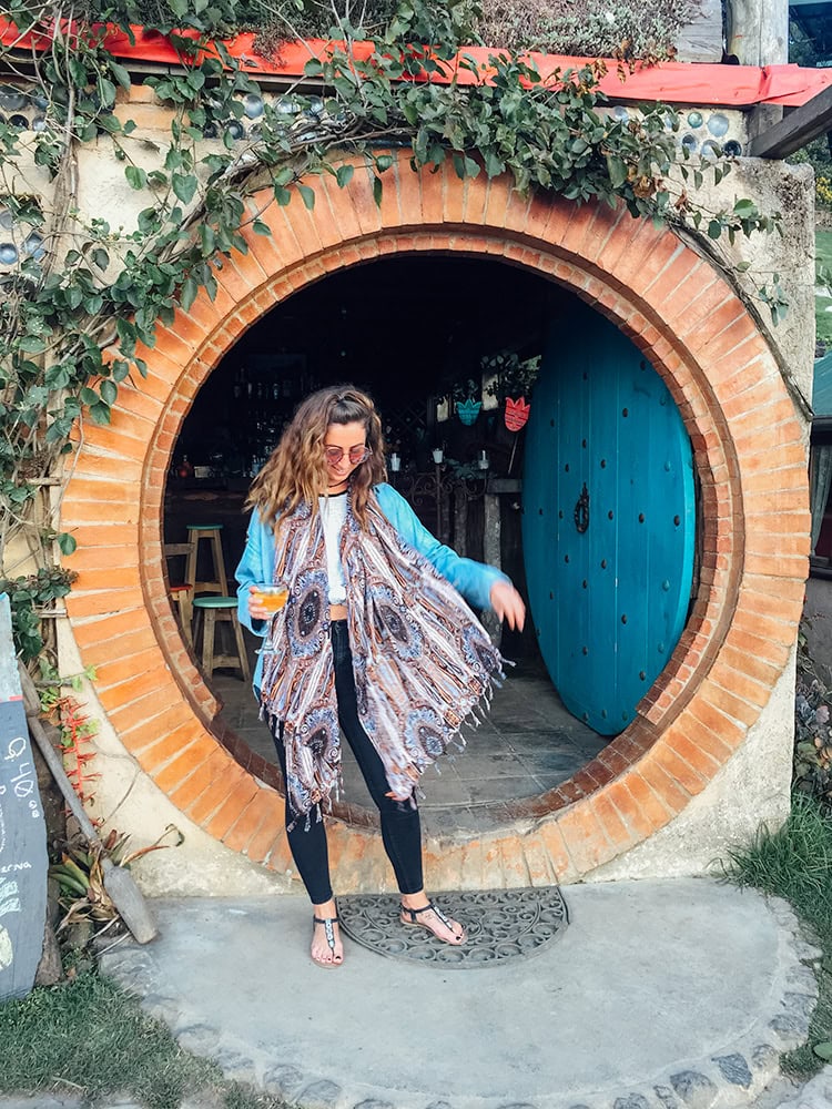 A woman wearing a flowing scarf stands in front of a circular brick doorway with a bright blue wooden door, leading into a cozy café. Hobbitenango, Guatemala