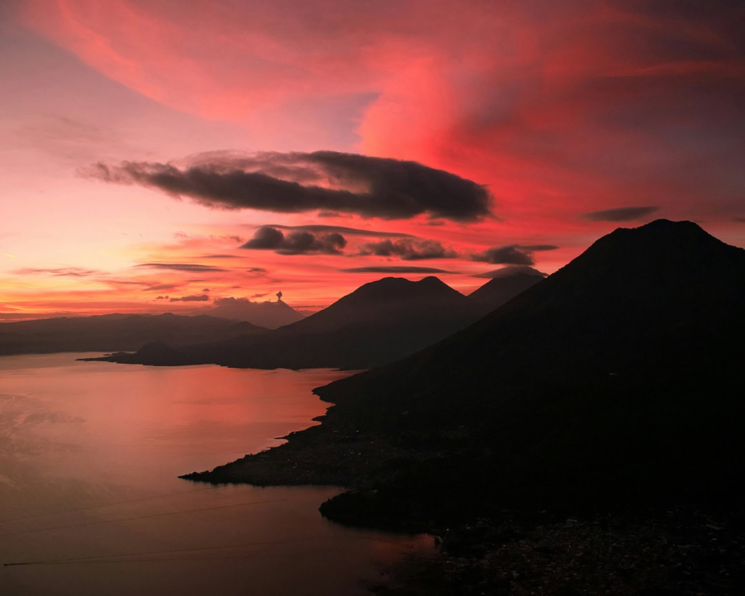 A fiery red and pink sunset lights up the sky over a lake, with dark volcanoes forming a striking silhouette. Indian Nose Hike, Lake Atitlán