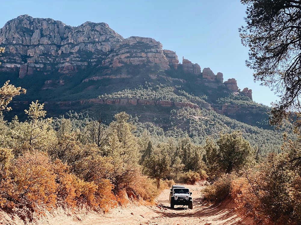 A rugged off-road vehicle driving on a dusty desert road, surrounded by red rock mountains and green vegetation.