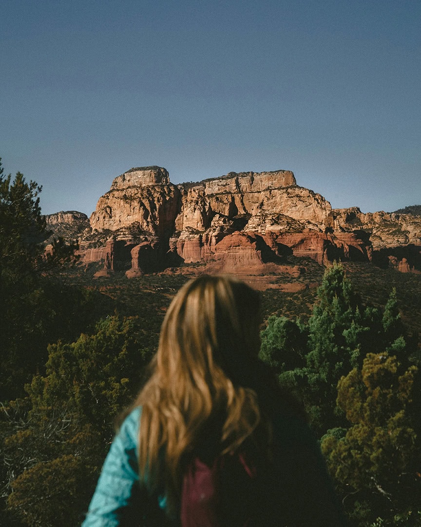 A woman sits on a red rock ledge, looking over a valley with towering red cliffs in the distance.
