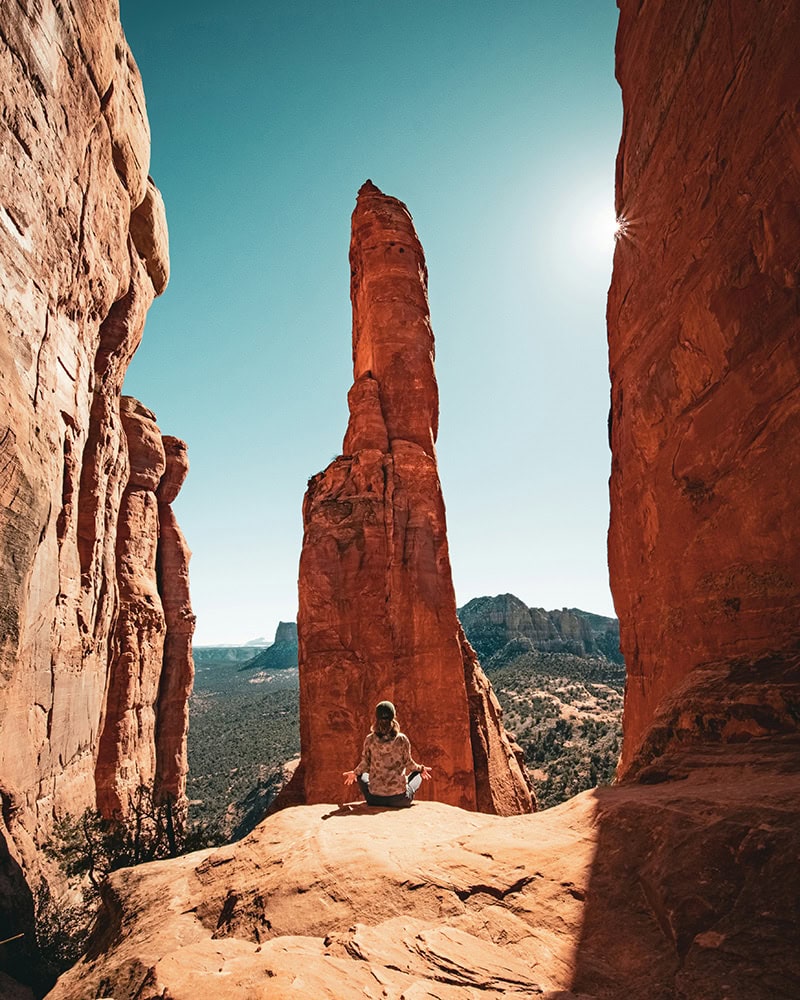 A woman sits cross-legged on a red rock ledge, meditating between towering rock formations.