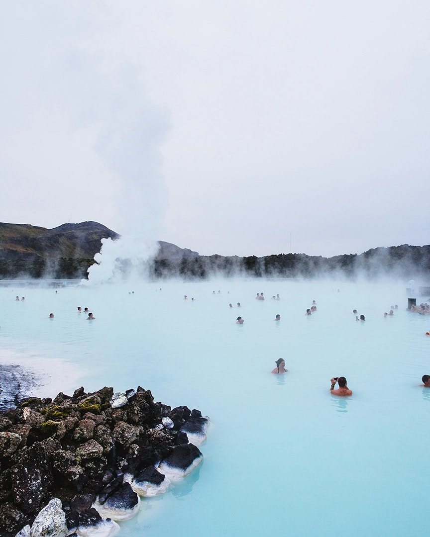 People relaxing in a large, milky-blue geothermal lagoon under a cloudy sky, with steam rising in the background.