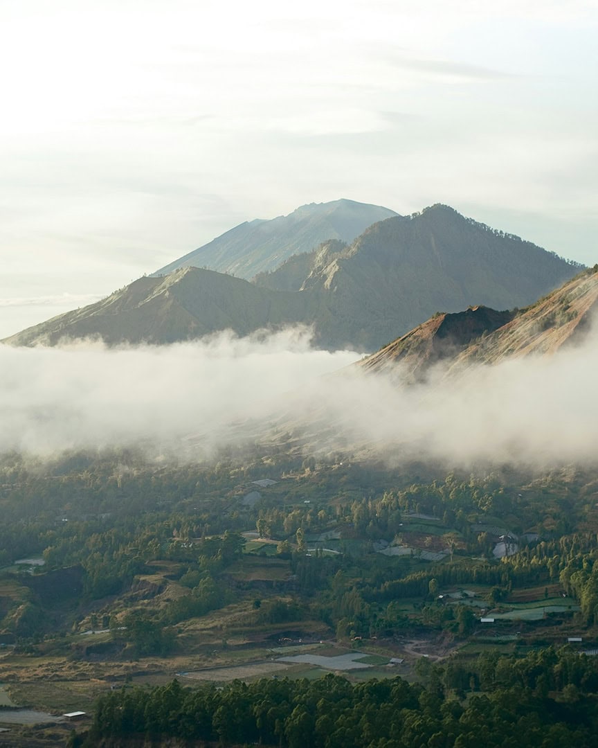 A misty view of a mountain range at sunrise, with soft clouds drifting through the valleys and patches of farmland visible below.
