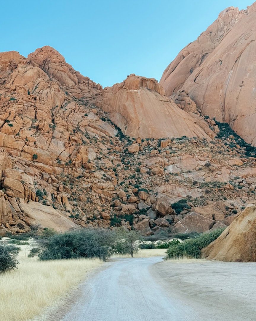 A dusty gravel road curves into a rugged landscape of massive reddish-orange granite mountains under a bright blue sky.